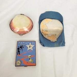 May include: Three vintage compacts are shown on a white surface. One is shell-shaped with pink and cream tones, another is silver with gold floral accents, and the third features a blue background with a film-themed illustration and the word "Paris".