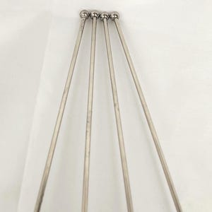 May include: Four silver metal cocktail skewers. Each skewer features a thin, straight shaft and a small, round ball at the top. The skewers are arranged in a fan shape against a white background.