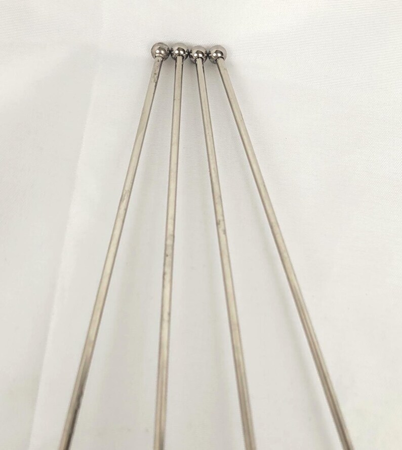 May include: Four silver metal cocktail skewers. Each skewer features a thin, straight shaft and a small, round ball at the top. The skewers are arranged in a fan shape against a white background.