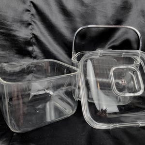 May include: A clear glass ice bucket with a metal handle and a square lid. The lid is partially open, revealing the interior. A separate square glass container is also present, suggesting a set for serving drinks.