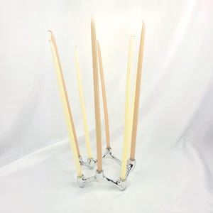 Peut inclure: Un bougeoir en métal argenté doté d'une structure interconnectée à plusieurs bras, supportant six bougies effilées hautes et fines. Les bougies sont de teintes alternées crème pâle et beige clair, dressées dans la base circulaire.