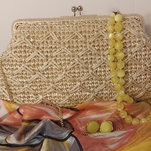 May include: A woven straw clutch bag with a gold clasp and a yellow beaded necklace draped over it. The bag is sitting on a colourful patterned fabric.