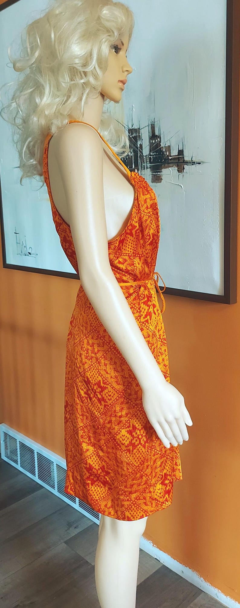 May include: An orange and red patterned dress with a halter neckline, displayed on a mannequin. The dress features a tie at the waist and a short hemline. The background includes an abstract painting and an orange wall.