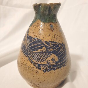 May include: A ceramic vase with a speckled tan body and a dark blue fish design. The top rim is green. The vase has a unique shape and a textured surface, showcasing a blend of rustic and artistic elements.