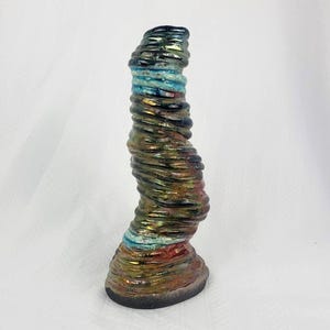 May include: A tall, vertical ceramic sculpture with a spiralling, ribbed texture. The surface features a metallic, iridescent glaze with blended tones of deep blue, copper, bronze, and turquoise, resting on a solid, dark, oval-shaped base.