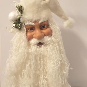 May include: A festive Santa Claus ornament with a cream-coloured hat and long, white beard. The face has realistic features with rosy cheeks and blue eyes. A small sprig of greenery and white balls adorn the hat. A holiday decoration.