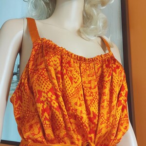 May include: A vibrant orange and red patterned dress with a tie waist and shoulder straps. The dress is displayed on a mannequin with blonde hair. The fabric features a repeating geometric design. The dress is a summer or resort style.