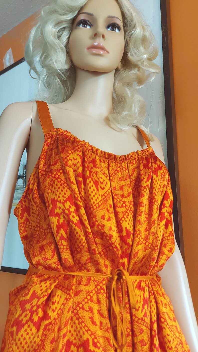 May include: A vibrant orange and red patterned dress with a tie waist and shoulder straps. The dress is displayed on a mannequin with blonde hair. The fabric features a repeating geometric design. The dress is a summer or resort style.