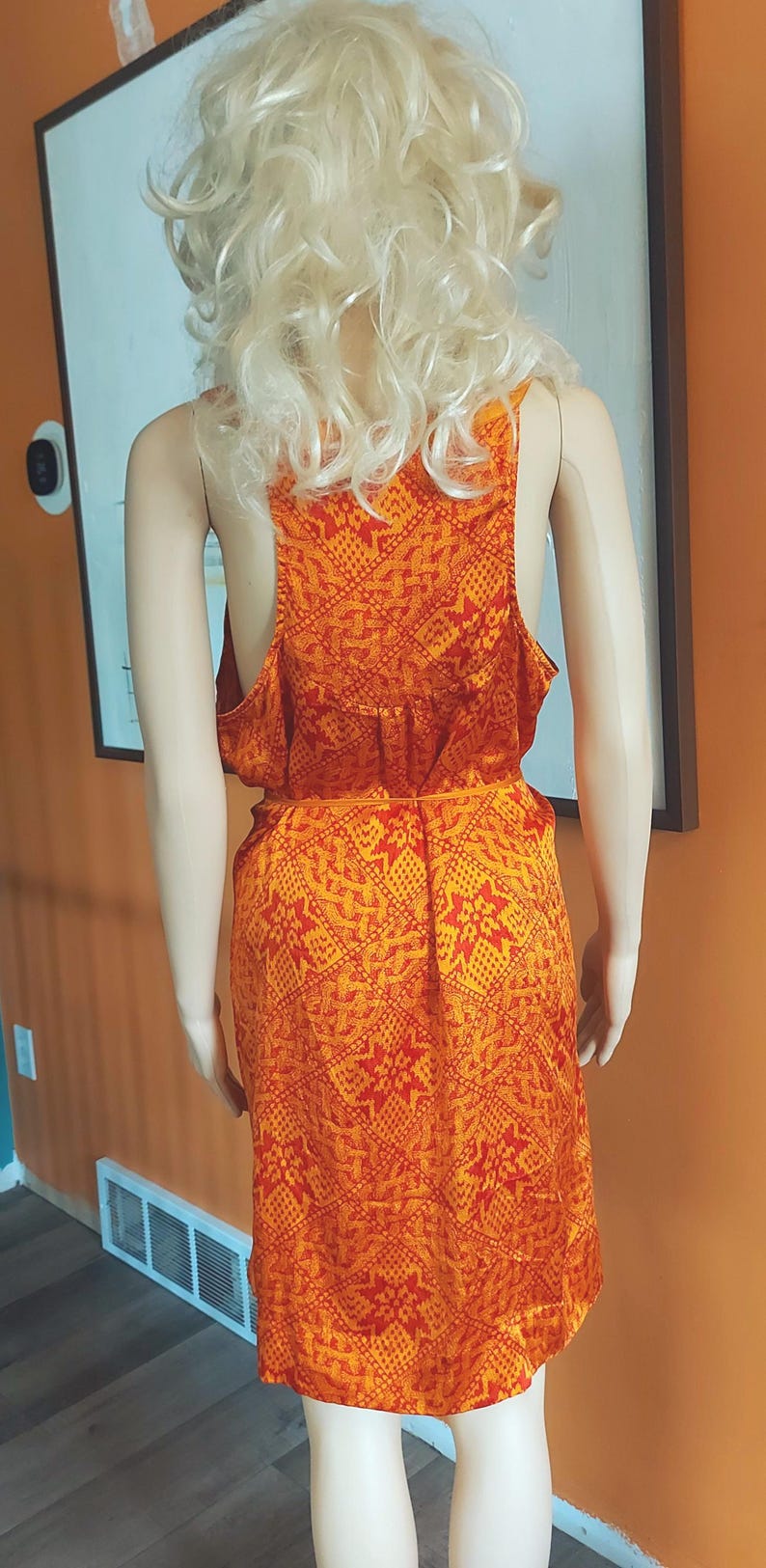 May include: A patterned orange and red dress on a mannequin. The dress features a sleeveless design, a scoop back, and a knee-length hemline. The dress is made from a lightweight fabric with a repeating geometric pattern.