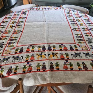 May include: A white linen tablecloth with a colourful border featuring a repeating pattern of people in traditional clothing, horses, and flowers.