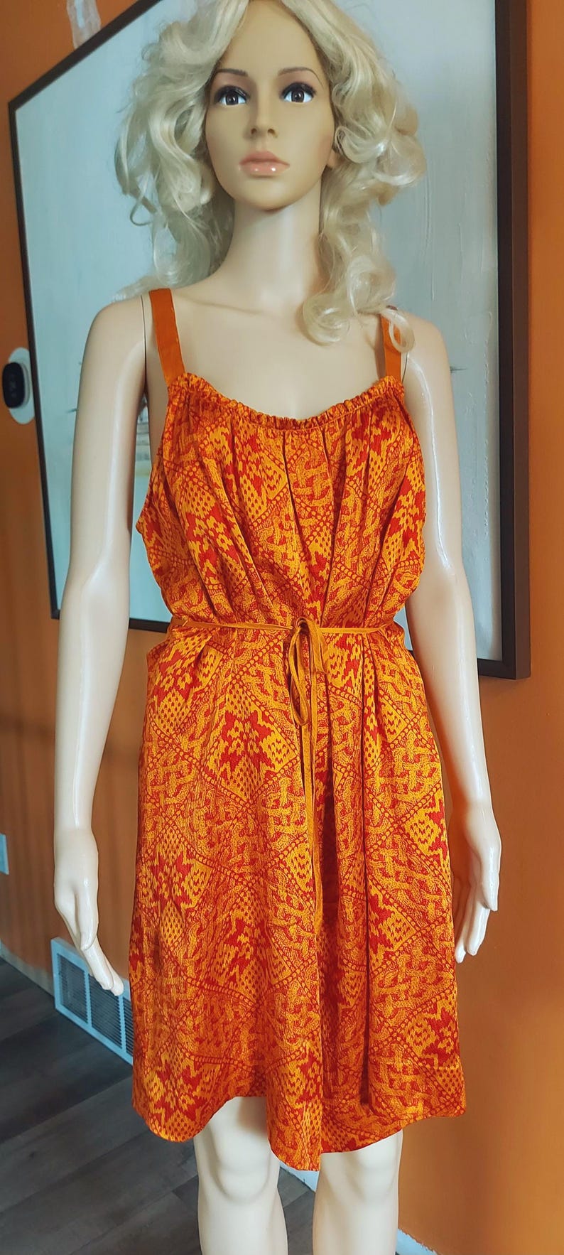 May include: Orange and red patterned dress with spaghetti straps. The dress features a tie waist and a repeating geometric design. The dress is displayed on a mannequin. The dress is approximately 140 cm long.