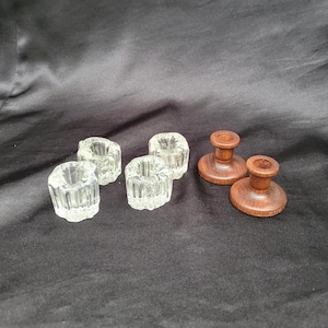 May include: A collection of six small candle holders consisting of four clear glass holders with a vertical ribbed texture and two smooth, circular brown wooden candle holders with a central pedestal and a wide, flat base for stability.