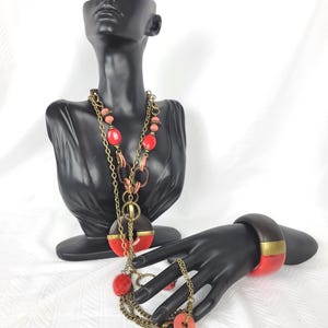 Peut inclure: Un long collier composé d'une chaîne en laiton, de perles rouges et corail, et d'éléments circulaires en bois brun foncé. Un bracelet jonc large assorti, avec une partie supérieure brune et une partie inférieure rouge, est présenté sur une main.