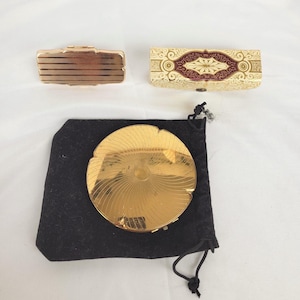 Peut inclure: Trois poudriers vintage et une pochette en velours noir sont présentés. Les poudriers comprennent un rectangulaire doré, un rectangulaire ivoire avec des accents rouges et un rond doré avec un motif radial, sur fond blanc.