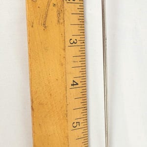 May include: A silver cocktail pick with a miniature top hat design. The pick is approximately 18 cm long, with a small ball at the bottom. A wooden ruler is in the background.