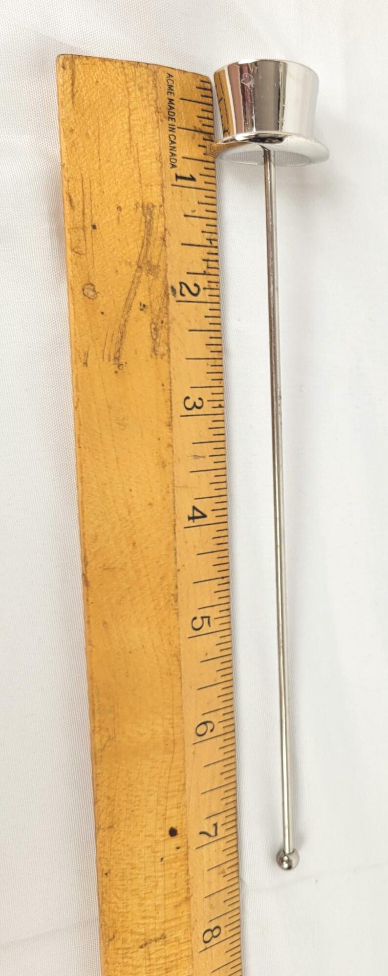 May include: A silver cocktail pick with a miniature top hat design. The pick is approximately 18 cm long, with a small ball at the bottom. A wooden ruler is in the background.