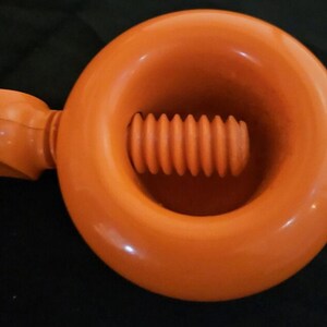 May include: An orange nutcracker with a circular design and a screw mechanism. The nutcracker has a handle for leverage and is made of a glossy plastic material. The tool is designed for cracking nuts.