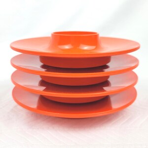 May include: A stack of four bright orange plastic egg cups. Each cup features a wide, flat circular base with a central raised holder designed to support an egg. The smooth, glossy finish of the orange material is consistent across all four pieces.