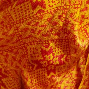 May include: Close-up of a fabric with a repeating pattern in shades of yellow and red. The design features geometric shapes and floral motifs, creating a textured appearance. The fabric appears to be soft and possibly silk or a similar material.