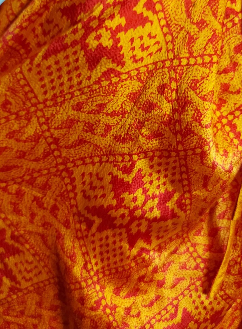 May include: Close-up of a fabric with a repeating pattern in shades of yellow and red. The design features geometric shapes and floral motifs, creating a textured appearance. The fabric appears to be soft and possibly silk or a similar material.