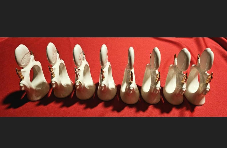 May include: Set of eight white ceramic napkin rings with gold accents. Each ring features a stylized angel design.