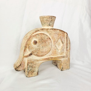 May include: A beige elephant-shaped decorative vase with a textured surface. The vase has a cylindrical neck and is carved with geometric and circular patterns. The elephant has four legs and a trunk. The vase is on a white background.