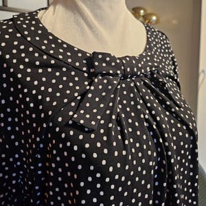 May include: A black blouse featuring a white polka dot pattern, a round neckline, and a decorative fabric bow detail centred at the chest. The garment includes vertical pleats extending downwards from the bow, creating a draped effect across the front.