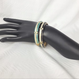 May include: Two hinged bangle bracelets featuring a glossy finish. One bracelet is turquoise and cream with gold-tone metal accents, while the other is black and cream with gold-tone metal accents. Both bangles have a smooth, rounded profile.