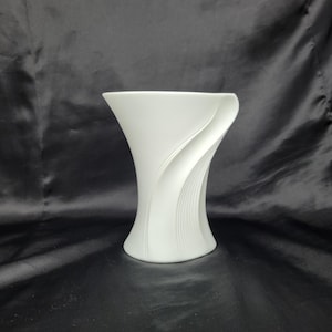 May include: A matte white ceramic vase featuring a smooth, flared top opening and a distinct, curved side panel decorated with a series of fine, vertical embossed lines that create a sense of fluid motion along the exterior surface.