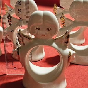 May include: Set of four white porcelain angel napkin rings with gold accents. Each ring features a small angel with wings and a halo. The angels are approximately 2.5 inches tall.
