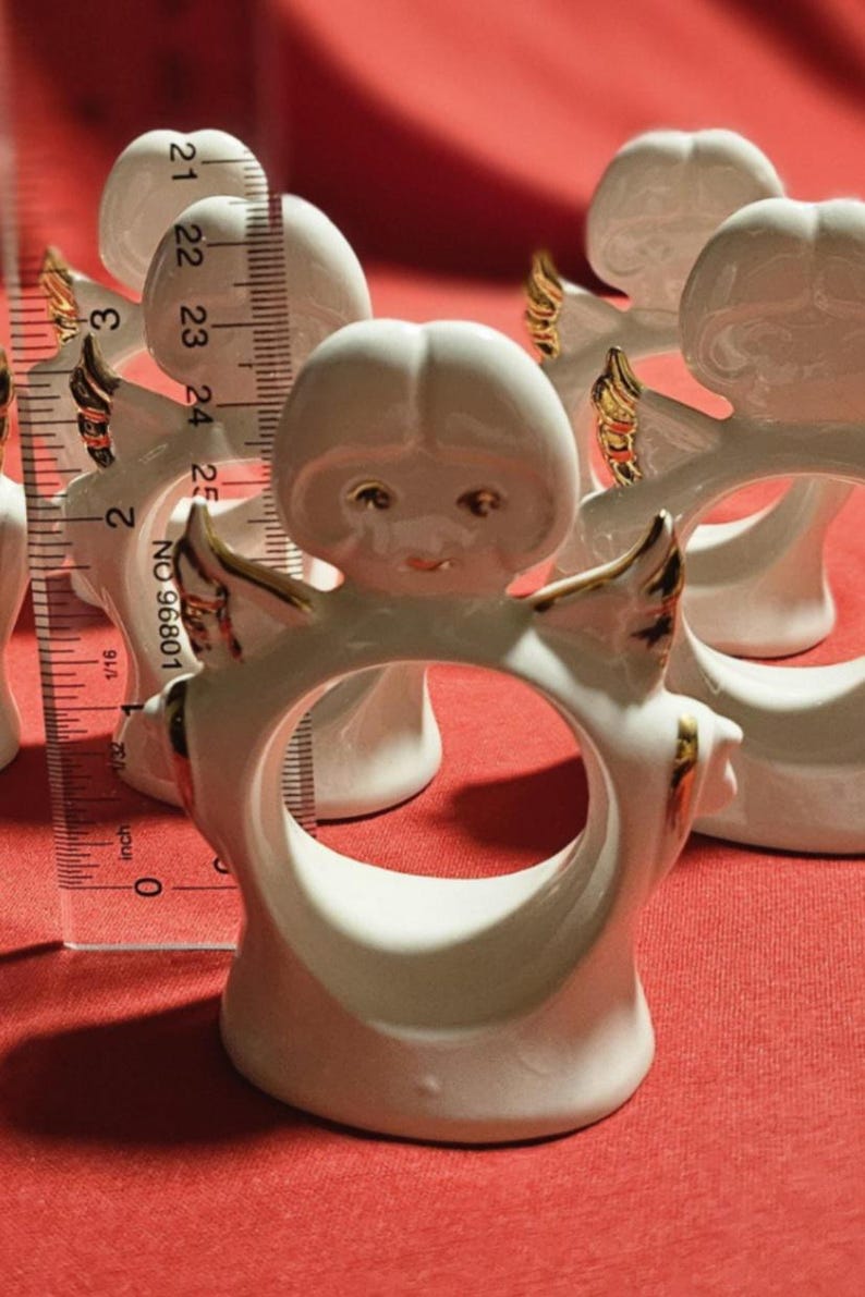 May include: Set of four white porcelain angel napkin rings with gold accents. Each ring features a small angel with wings and a halo. The angels are approximately 2.5 inches tall.