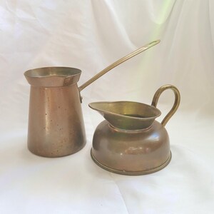 May include: A metallic copper-toned coffee pot with a long handle and a matching metallic copper-toned creamer pitcher featuring a curved handle and a wide pouring spout, both displaying a smooth, polished finish and a cylindrical base.