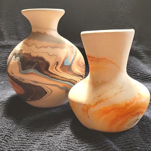 May include: Two ceramic vases with a marbled design. One vase features swirls of brown, black, and blue. The other vase has orange and white marbling. Both vases have a light beige colour at the top.