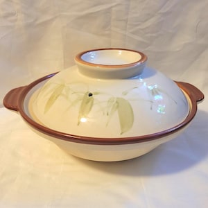 May include: Cream-colored ceramic Japanese donabe pot with a lid. The pot features a brown rim and handles, with a bamboo leaf design in green. The lid has a small opening and a brown rim.