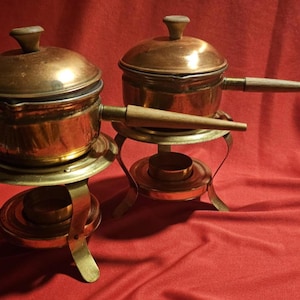 Peut inclure: Deux casseroles en cuivre avec couvercles, chacune dotée d'un long manche en bois et d'un couvercle en cuivre assorti avec un bouton en bois. Les casseroles reposent sur des supports en laiton, chacun équipé d'une base de brûleur circulaire.