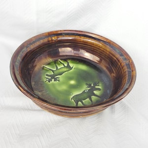 May include: A round ceramic bowl featuring a glossy brown exterior and rim. The interior base is a vibrant lime green colour, decorated with two dark silhouettes of moose standing in opposite directions, finished with a smooth, reflective glaze.