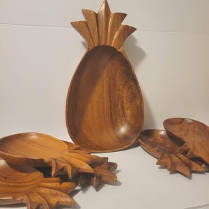 May include: Wooden pineapple-shaped serving bowls and plates. The large bowl is shaped like a pineapple with a deep interior. Smaller plates are shaped like pineapple tops. The wood has a warm, natural finish.