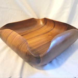 May include: A square wooden bowl with gently curved sides. The bowl is made of a light brown wood with darker brown grain patterns. The edges of the bowl are smooth and rounded. The bowl is empty and sits on a white surface.