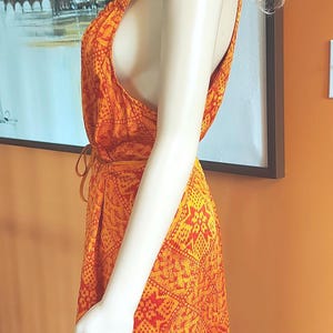 May include: An orange and red patterned dress with a halter neckline. The dress is sleeveless and has a tie at the waist. The dress is on a mannequin. The background is an orange wall and a painting.