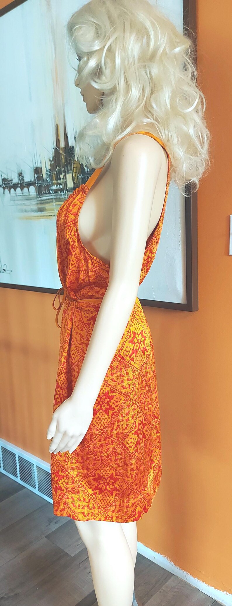 May include: An orange and red patterned dress with a halter neckline. The dress is sleeveless and has a tie at the waist. The dress is on a mannequin. The background is an orange wall and a painting.