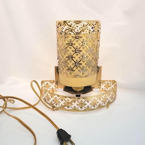 May include: A vintage gold-toned wall sconce with a decorative, openwork design. The lamp features a cylindrical shade and a matching base with a floral pattern. A brown electrical cord and plug are visible.