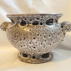 May include: A ceramic vase with a speckled, textured surface in shades of gray and brown. The vase has a wide, open top with two small handles and a pedestal base. The design features a pattern of small, dark brown spots.