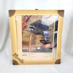 May include: A framed embroidery kit featuring a raccoon, a rifle, and a kettle. The kit is labeled "Bandit" and measures 12" x 13". The frame is light brown with dark brown corner accents. The brand name "afronelle" is visible.
