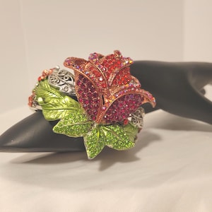 May include: A floral-themed bracelet featuring a large, ornate rose. The rose is composed of red and pink crystals, with green crystal-studded leaves. The bracelet has a silver-colored filigree design and is set on a black display.