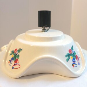 Peut inclure: Support de sapin de Noël en céramique crème avec un dessus en plastique noir. Le support présente un bord ondulé et est décoré de cloches colorées et de motifs de brins de conifères. Une vis métallique fixe le dessus noir au support.