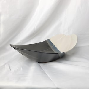 May include: A ceramic serving bowl with a unique design. The bowl is primarily dark grey, transitioning to white with a blue accent. The bowl has a curved, boat-like shape, perfect for serving salads or displaying fruit.