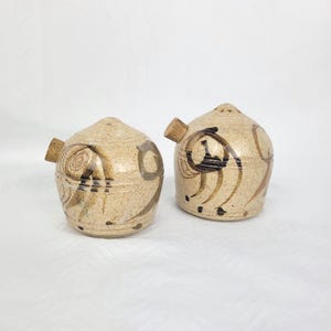 May include: Two round, speckled beige ceramic shakers with a dome-shaped top and a small cork stopper on the side. Each piece features dark brown, hand-painted abstract swirling patterns and lines across the textured, light-coloured clay surface.