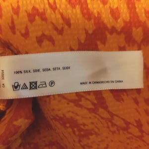 May include: A white label with text reading "100% SILK, SOIE, SEDA, SETA, SEIDE" and "MADE IN CHINA/HECHO EN CHINA". The label is set against a vibrant orange and red patterned fabric.