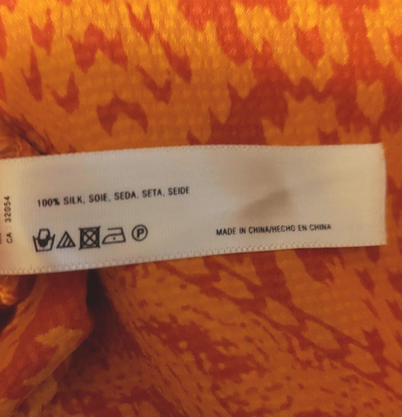 May include: A white label with text reading "100% SILK, SOIE, SEDA, SETA, SEIDE" and "MADE IN CHINA/HECHO EN CHINA". The label is set against a vibrant orange and red patterned fabric.