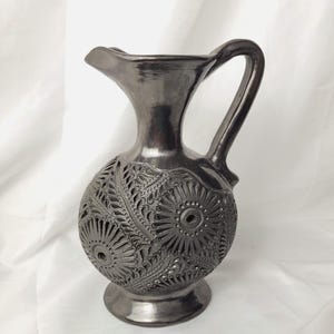 May include: A dark grey ceramic pitcher with a glossy finish, featuring a rounded body adorned with intricate, carved floral and sunburst patterns. The vessel has a narrow neck, a flared rim, a curved handle, and a circular pedestal base.